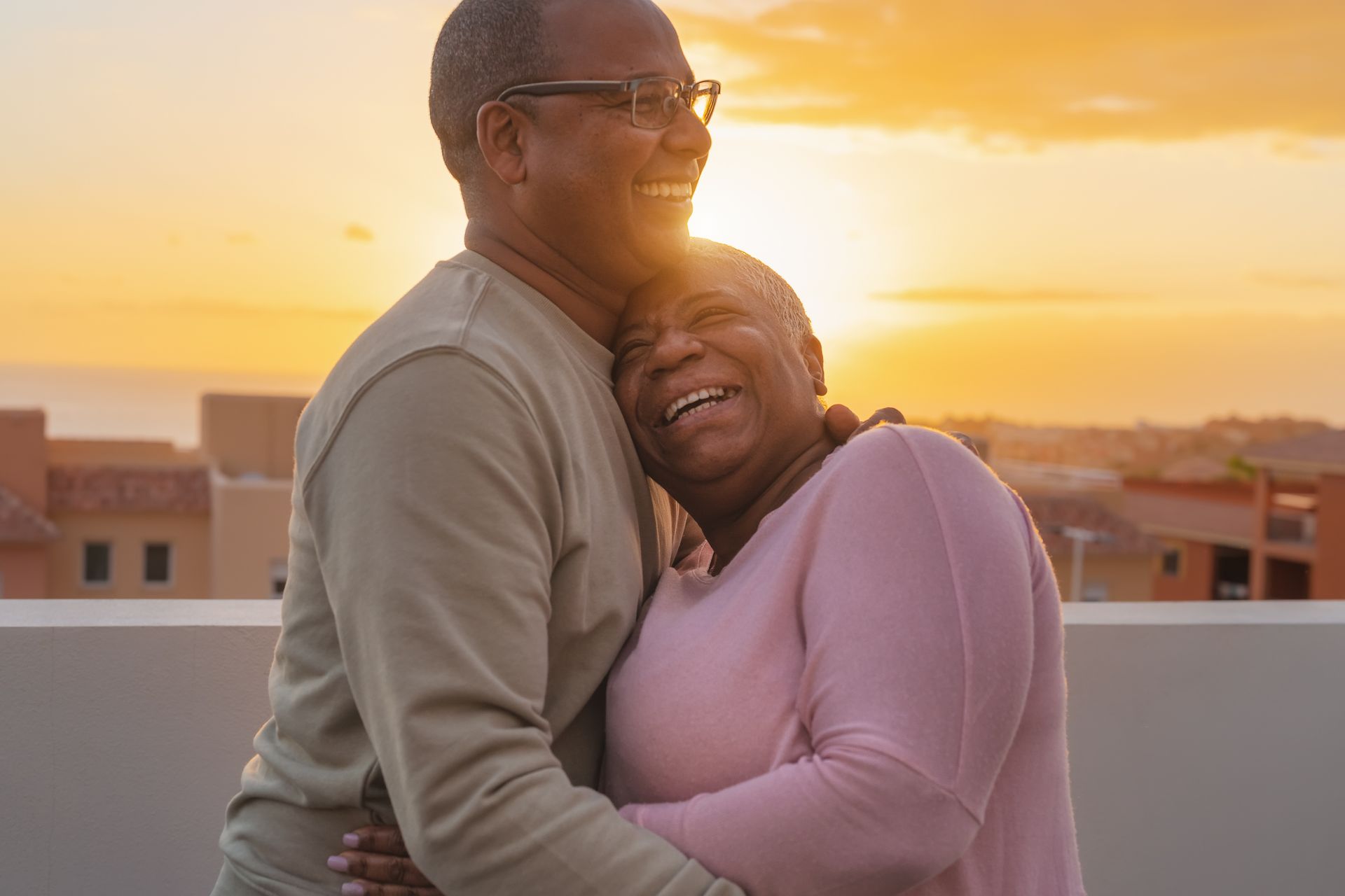 Two people embrace on a rooftop at sunset, smiling warmly with the sun glowing behind them.