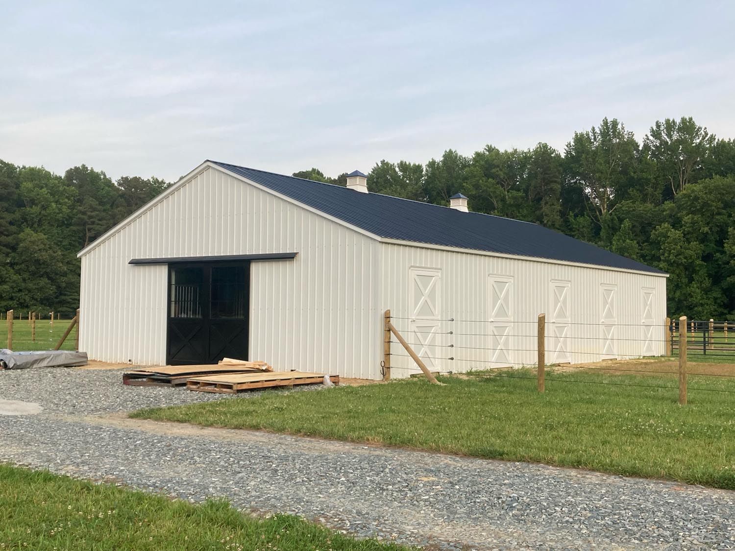 A large white barn with a black roof is sitting in the middle of a grassy field.
