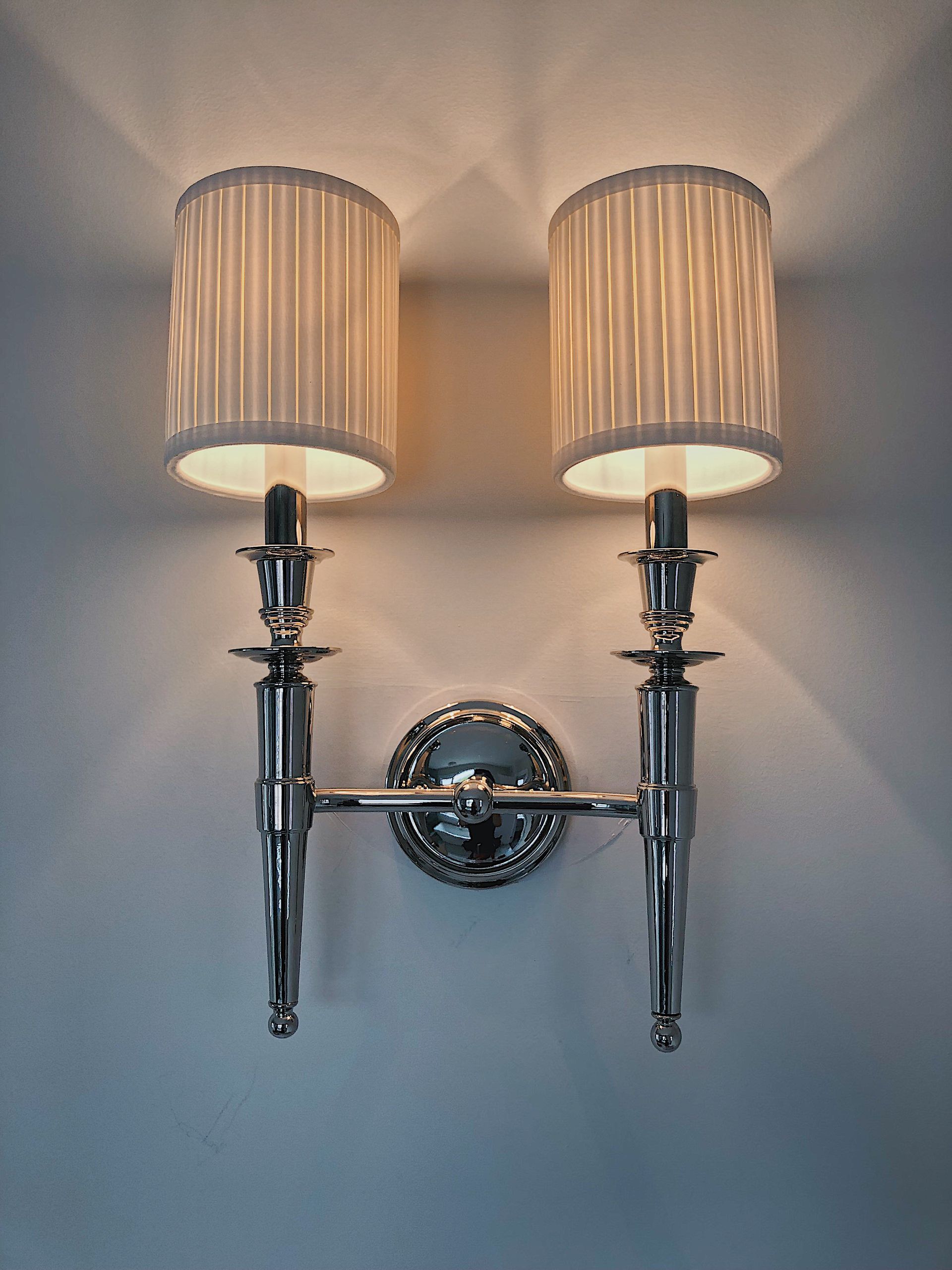 A polished chrome double-arm wall sconce with two pleated, off-white cylindrical fabric shades, mounted on a white wall.