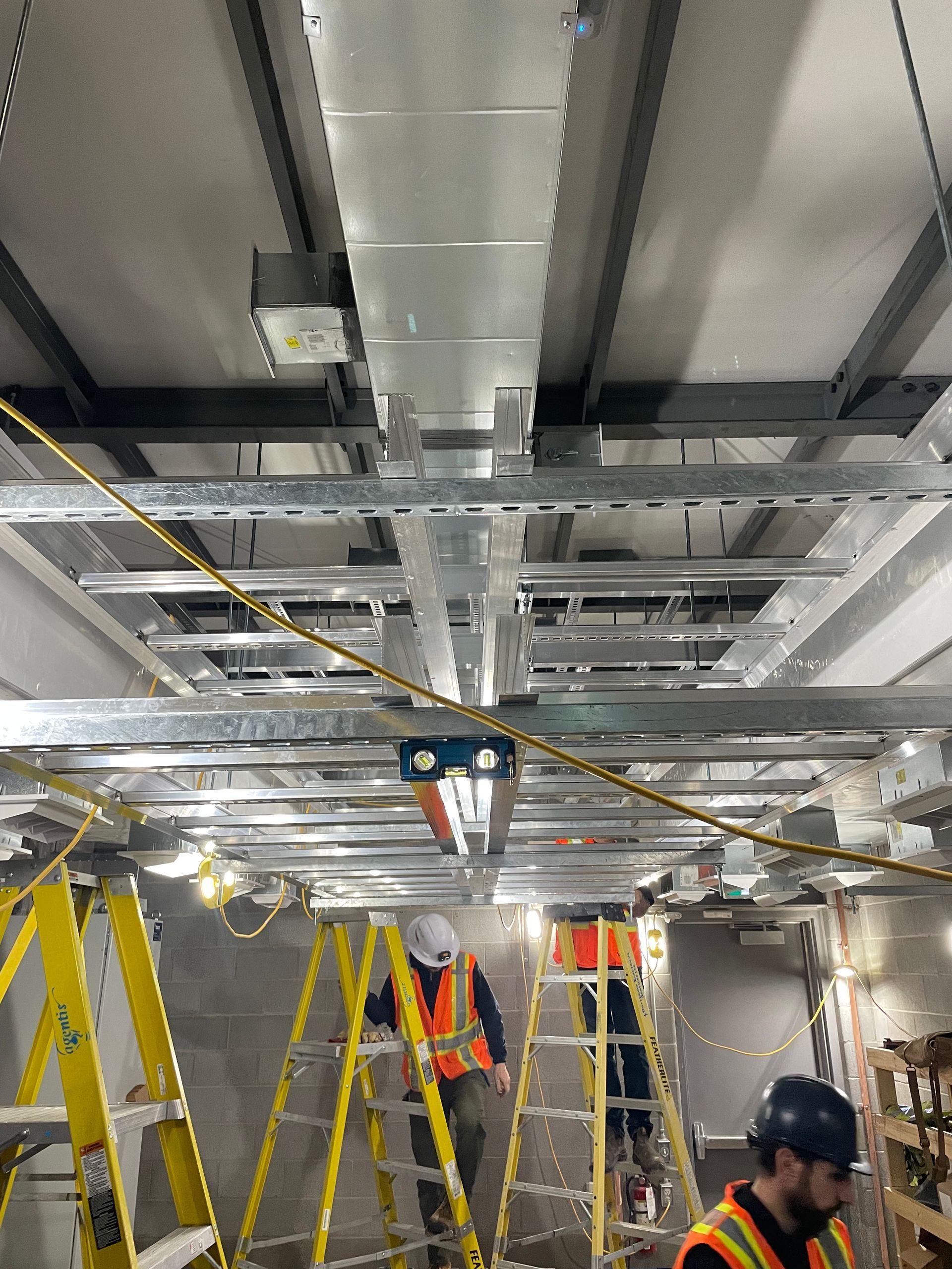 Construction workers on yellow ladders in a metal-framed ceiling space, one worker wearing a hard hat.