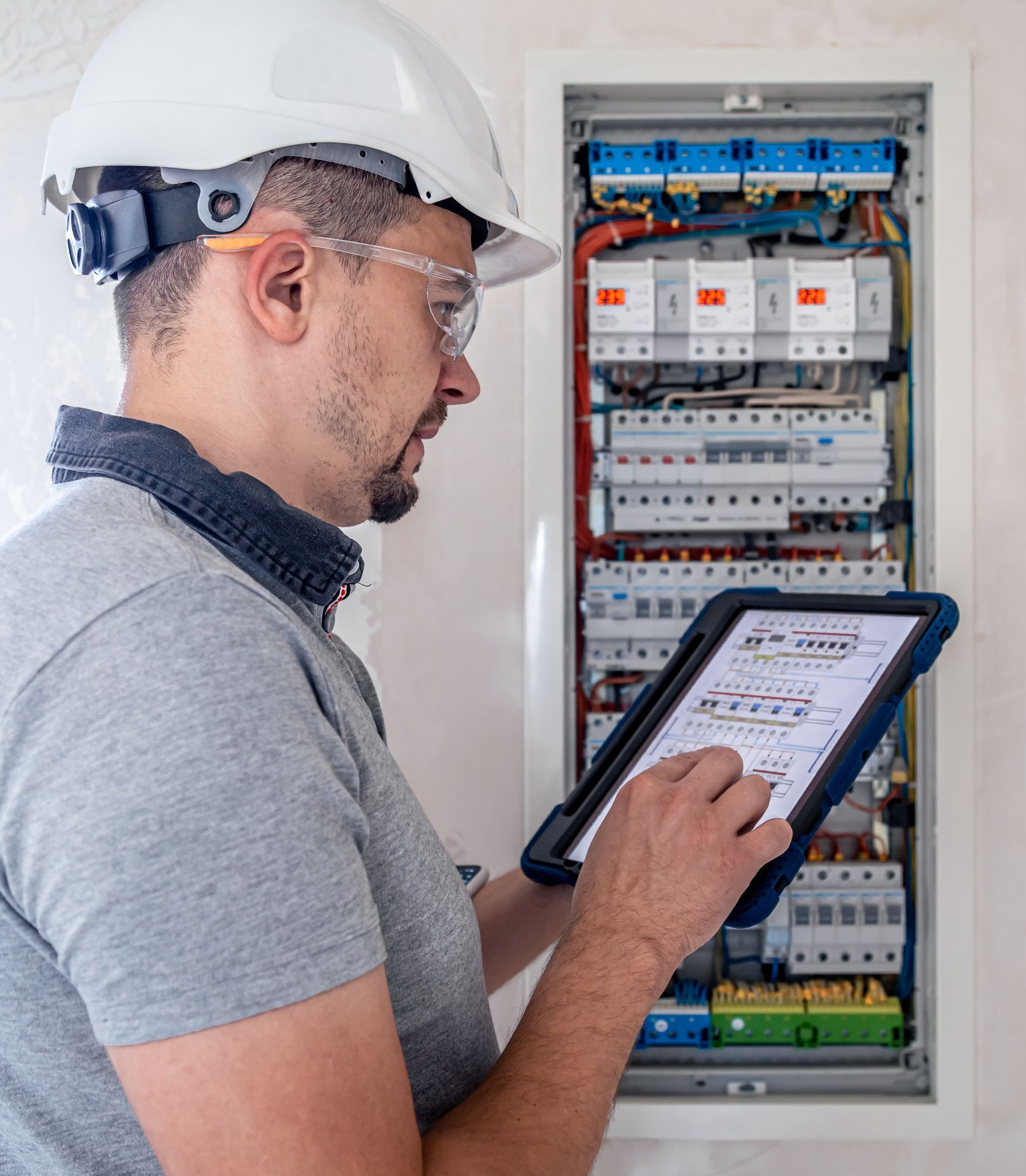 A man in a hard hat is looking at a tablet in front of an electrical box.