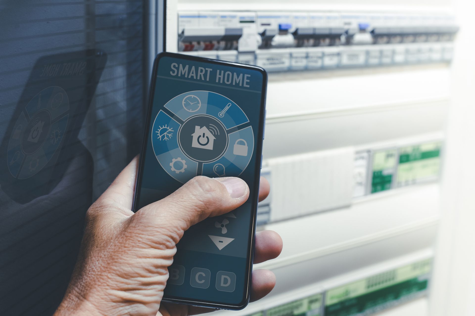 Hand holding a smartphone displaying a smart home control interface, in front of a breaker box.