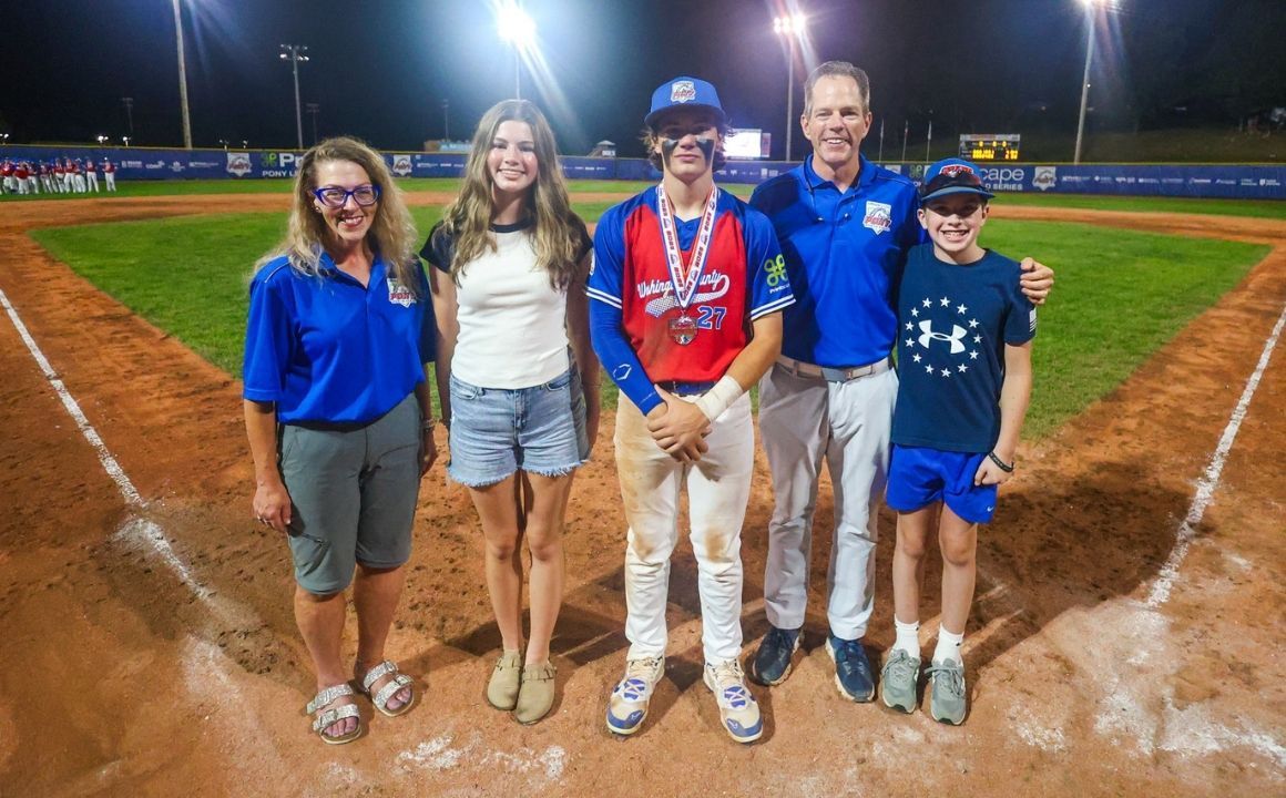 Five people pose on a baseball field at night near home plate under bright stadium lights.