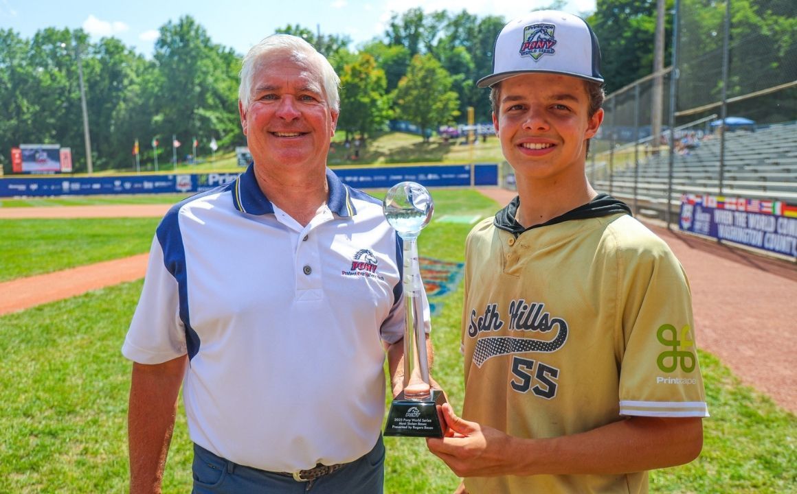 Two men on a baseball field holding a trophy, smiling in front of the stands and diamond.