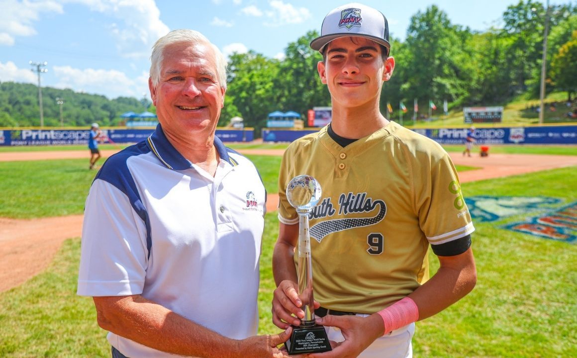 Two men on a baseball field hold a trophy, smiling in front of the outfield.