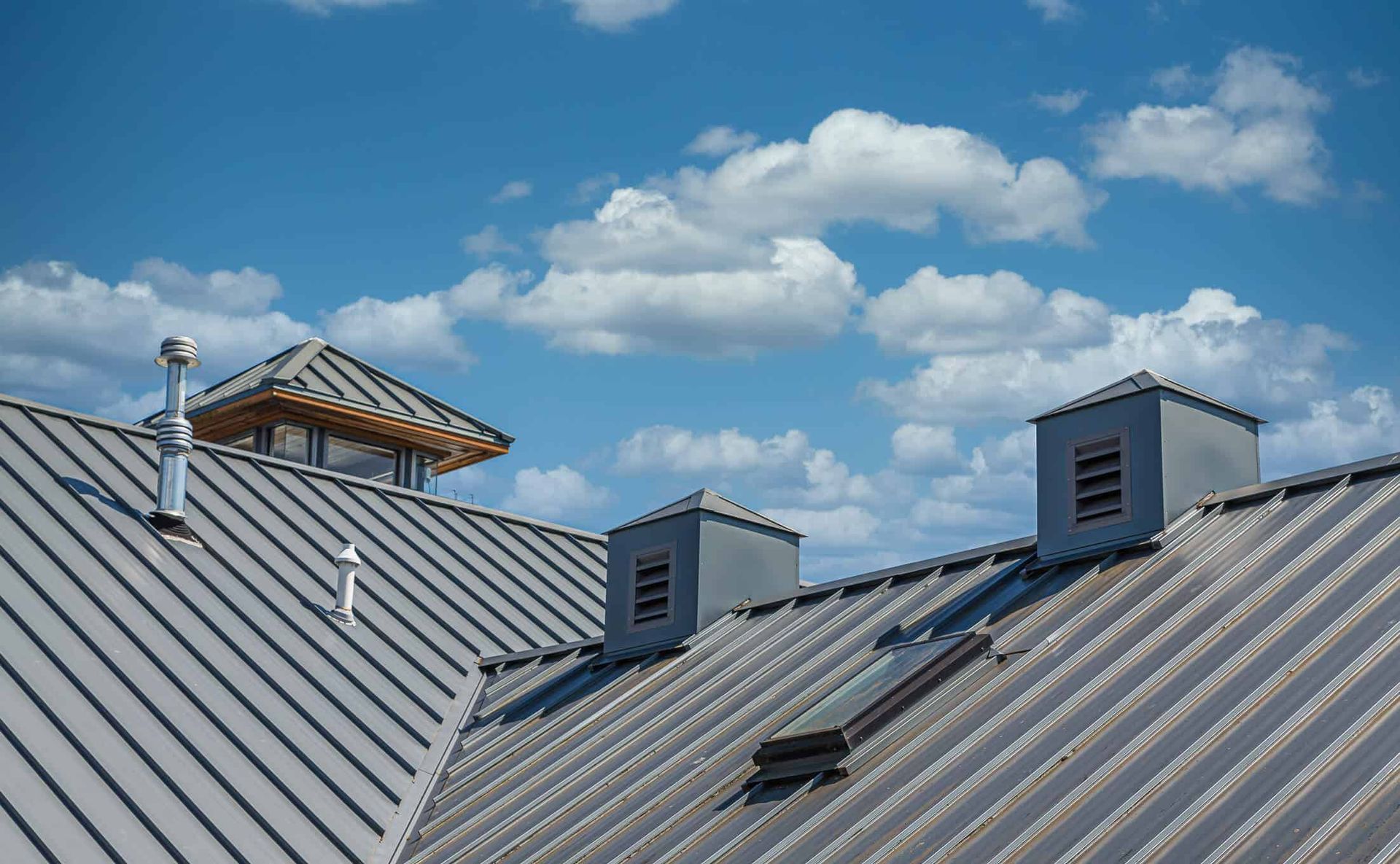 Metal roof with vents and a cupola against a blue sky with clouds.