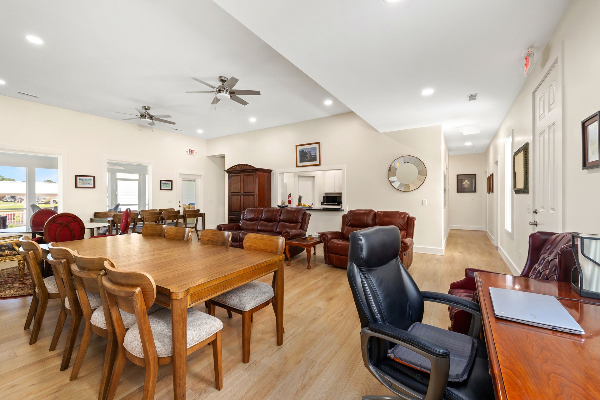 Spacious, light-filled room with a dining table, seating area, and office setup; neutral walls, wood floors, and various furnishings.