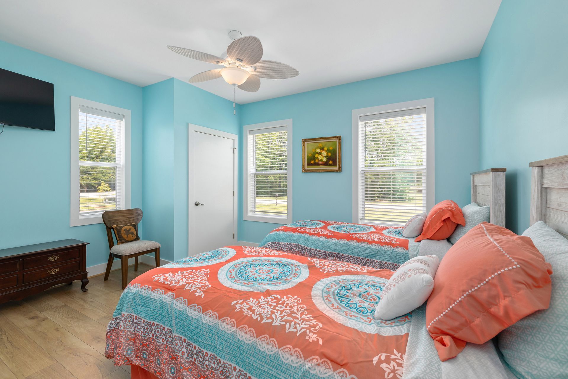 A bedroom with two beds, teal walls, and wood flooring. The beds have orange and teal bedding, and there's a tv, dresser, and chair.