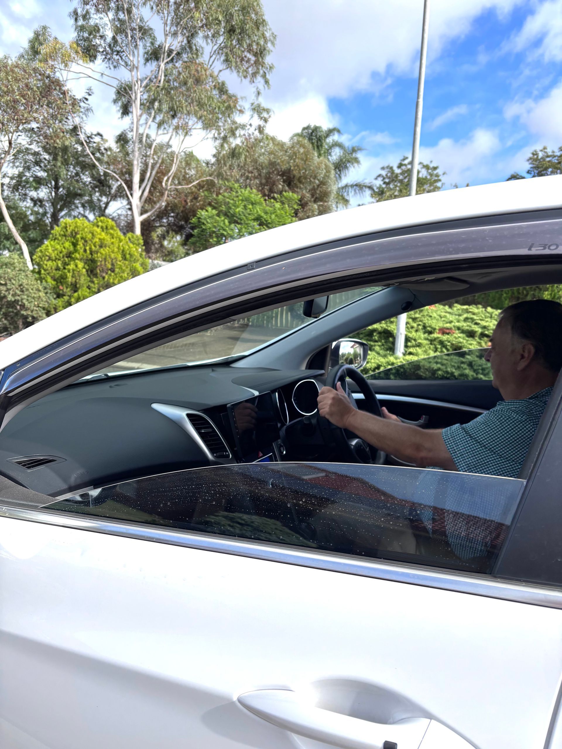 Man driving a silver car, seen through the side window on a sunny road.