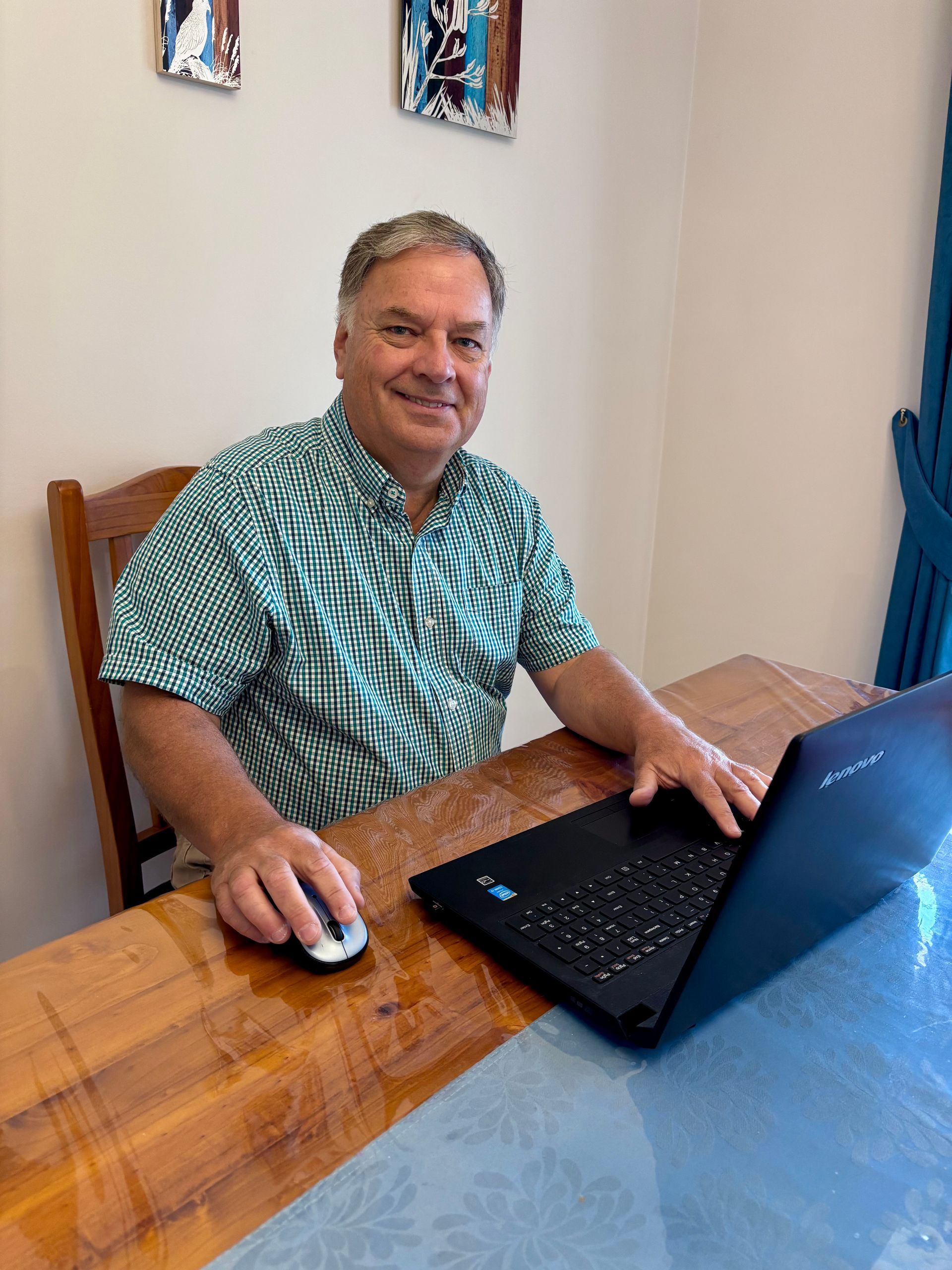 Man seated at a wooden table using a laptop and mouse, smiling in a small home office.
