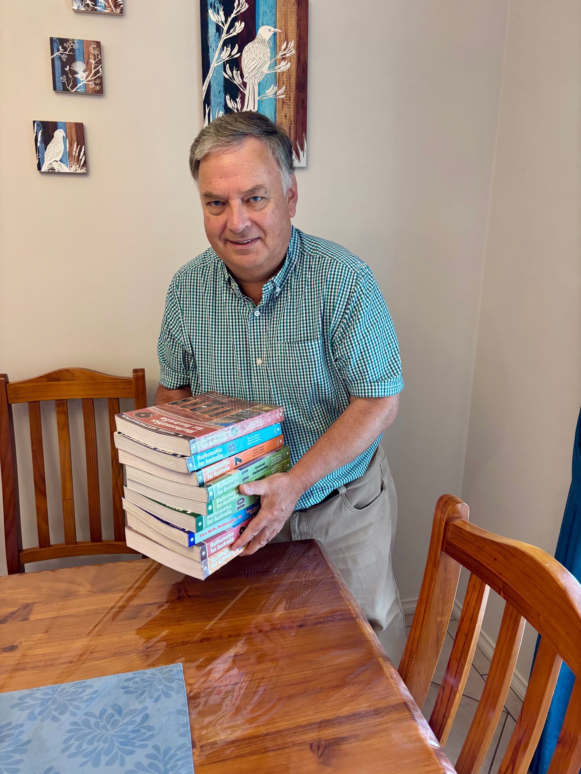 Man holding a stack of books beside a wooden table and chairs on a white background