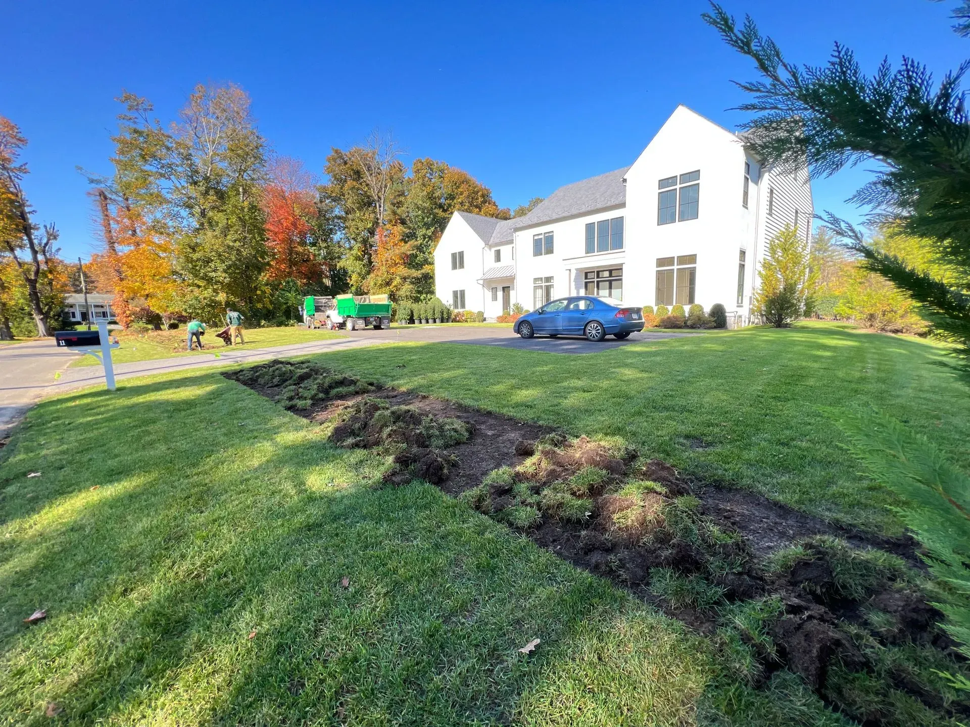 Lawn being dug up in front of a white house. Blue car parked in driveway, trees in background.