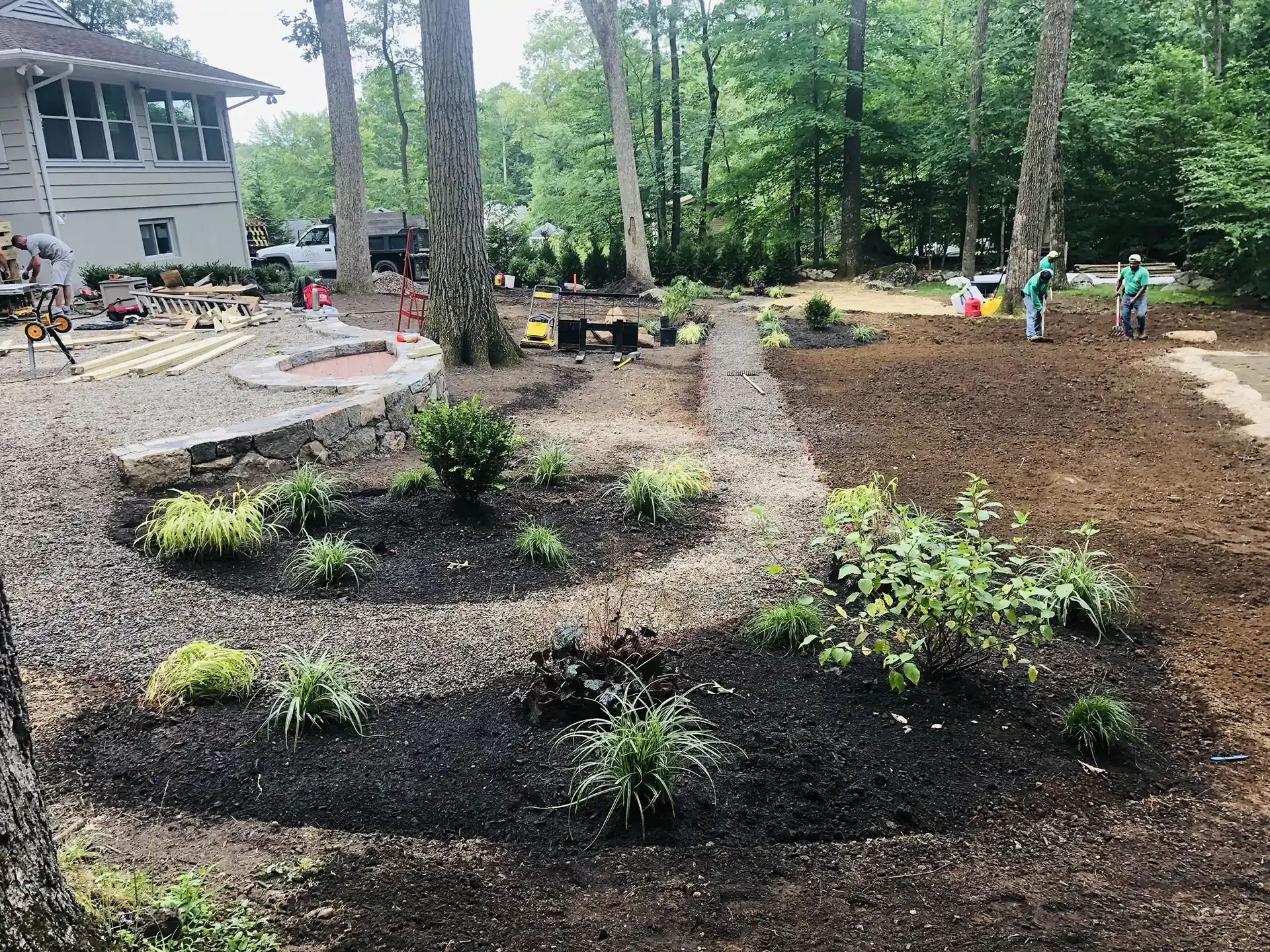 Landscaped yard with mulch, plants, stone path, fire pit, and workers in background.