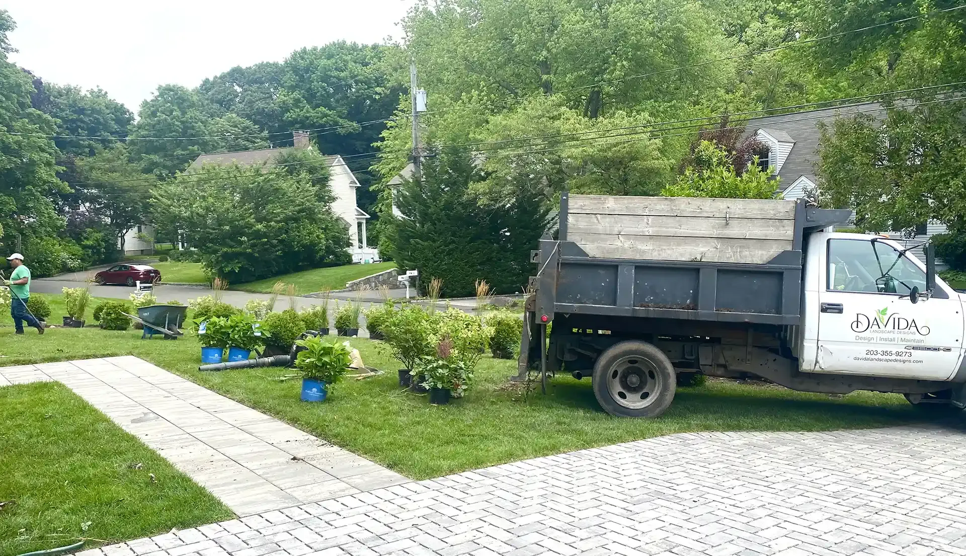 Landscaping truck parked on grass, next to a sidewalk, with a person planting bushes.