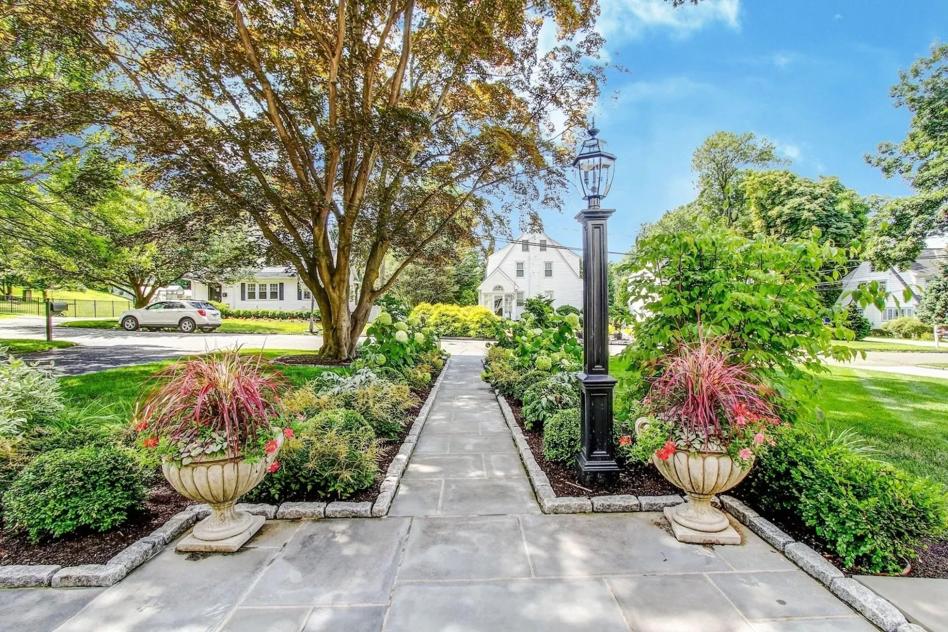 Stone pathway leading to a white house, lined with flowerbeds and planters, under a big tree on a sunny day.