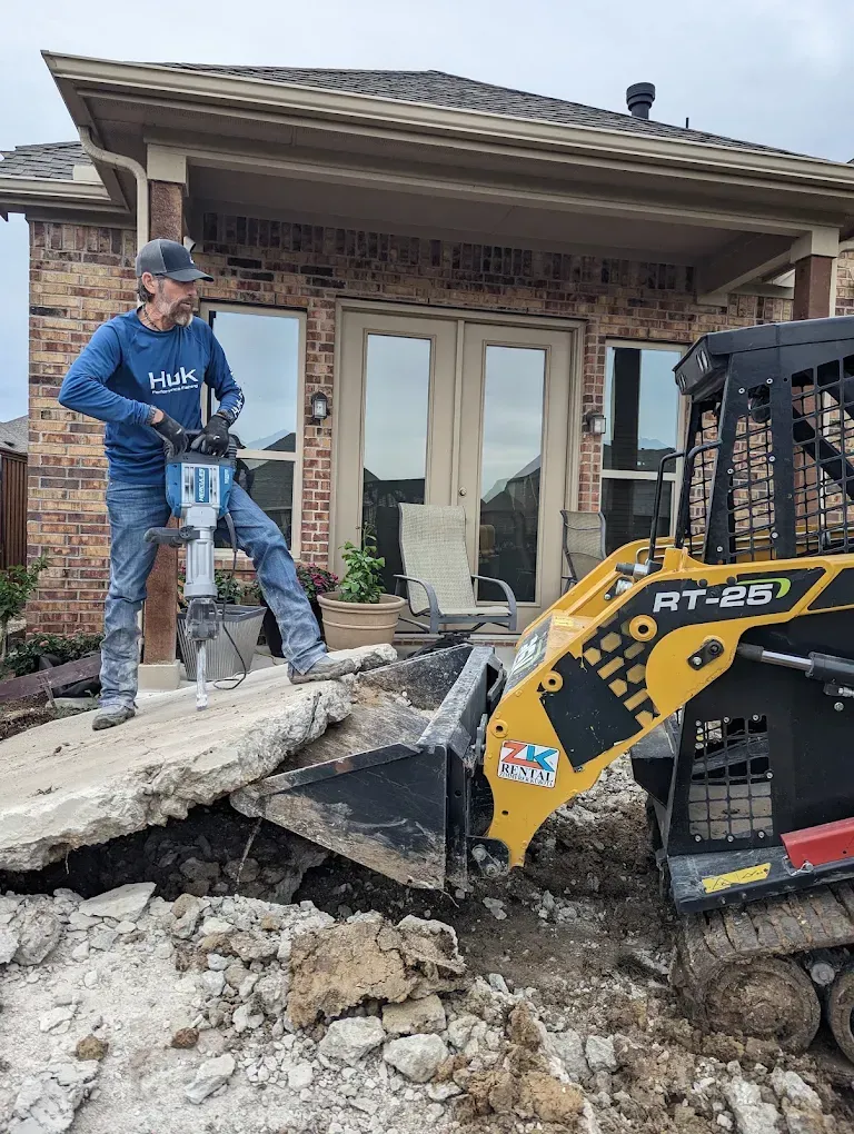 Worker using a jackhammer beside a construction loader in front of a brick house with a damaged porch.