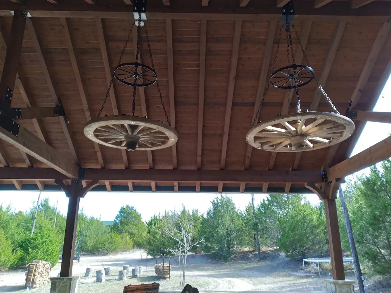 Wooden pavilion roof with two hanging wagon wheels above an open sandy outdoor area and trees beyond