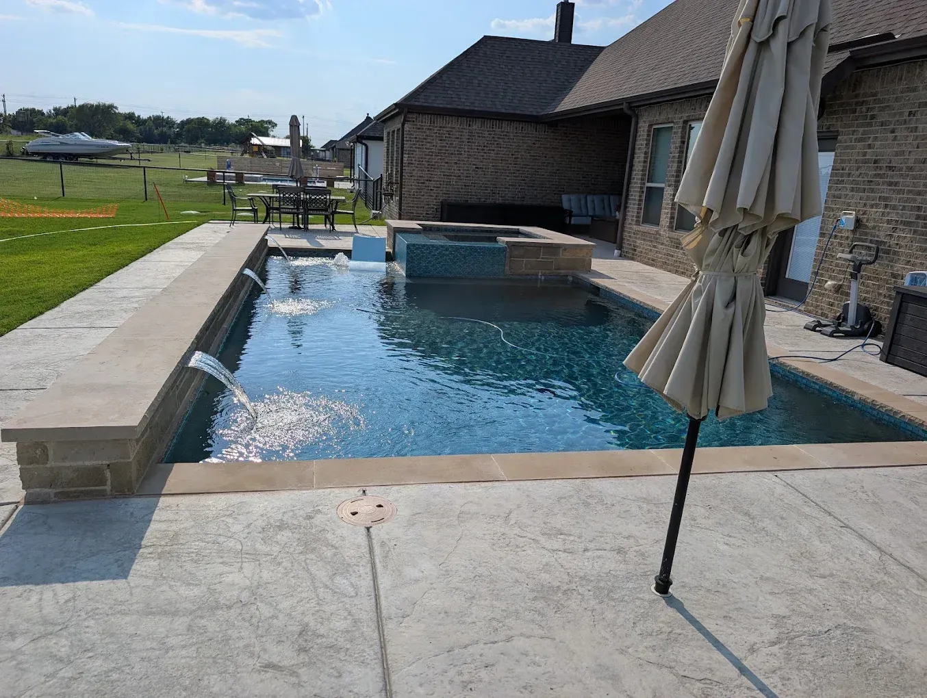 Backyard swimming pool beside a house, with a patio and closed umbrellas on a sunny day