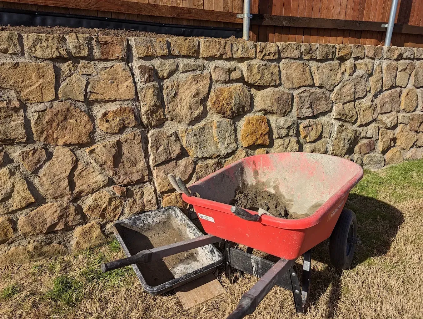 Red wheelbarrow and black shovel with dirt beside a stone wall in a yard