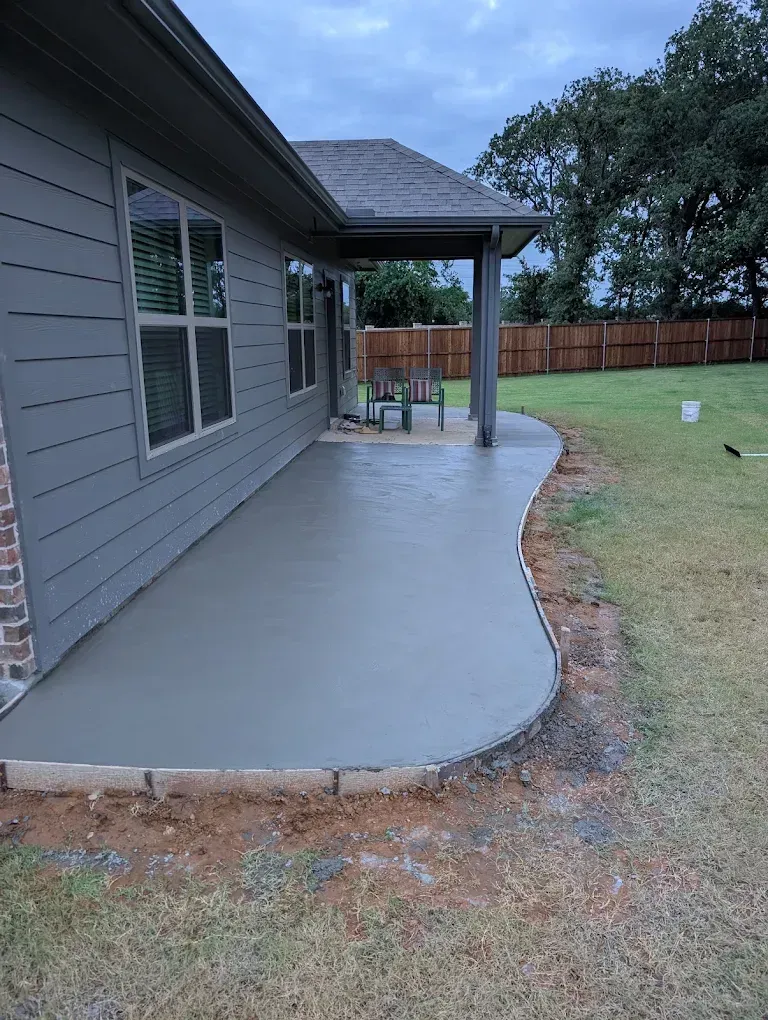 Concrete patio along gray house with covered porch and curved edge beside a grassy yard.