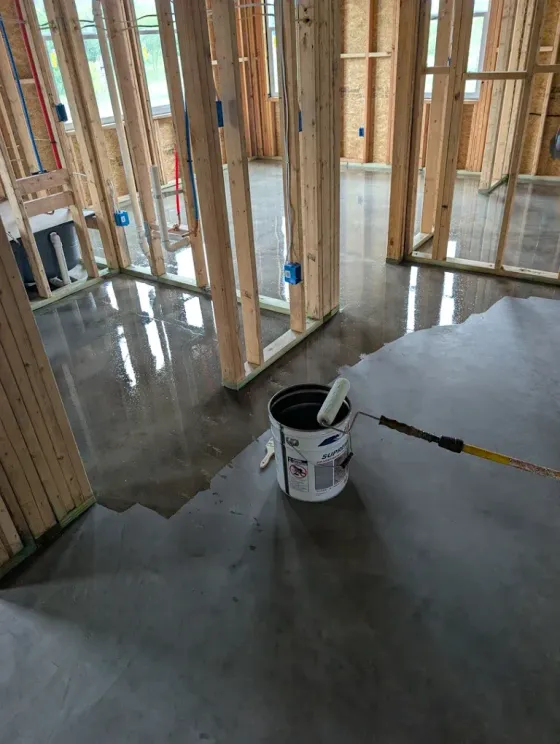 Unfinished house interior with framed walls, wet concrete floor, and a bucket with a trowel in the foreground