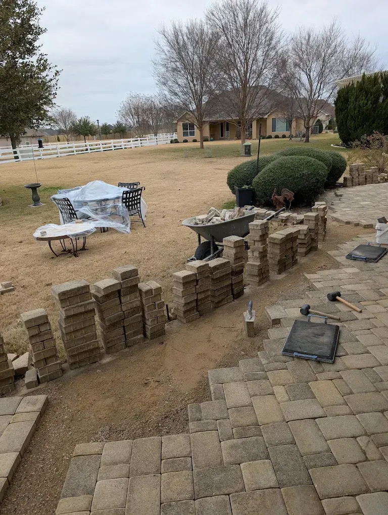 Backyard patio with stacked pavers, a small bench, and leafless trees under a cloudy sky