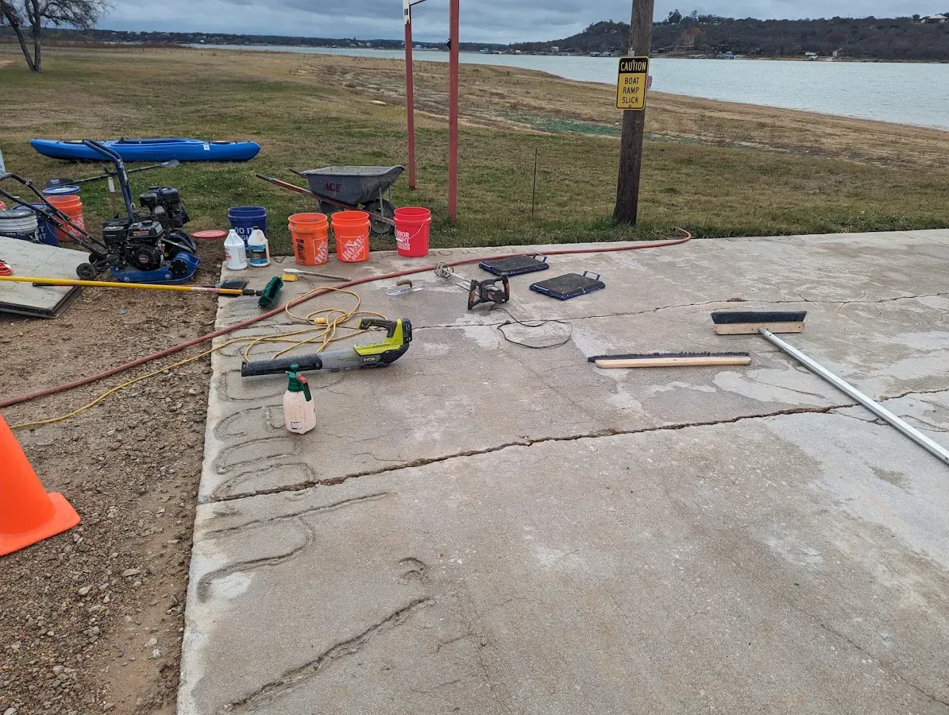 Outdoor work area with tools, cones, and buckets on concrete beside a grassy waterfront.
