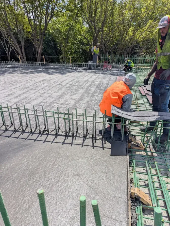 Workers installing rebar on a concrete slab at a construction site, one in an orange jacket bending over.