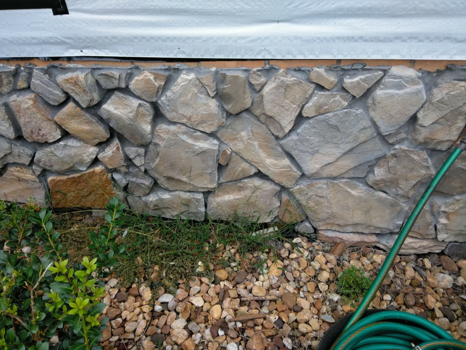 Stone foundation wall with gravel and a green garden hose at the base
