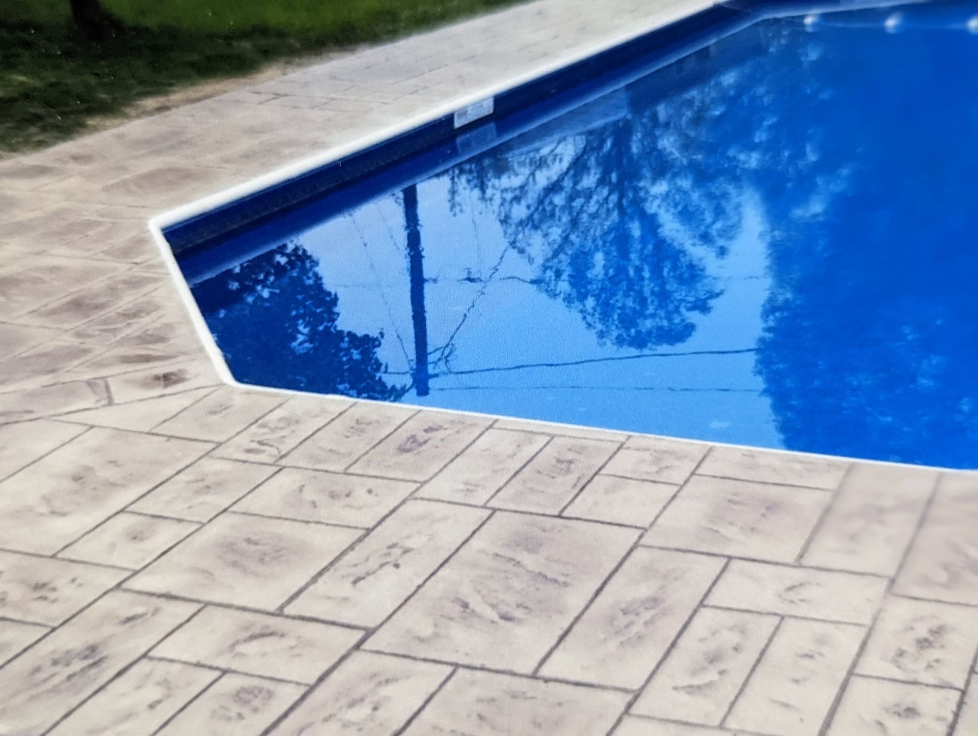 Blue swimming pool beside a light stone patio, with trees reflected in the water.