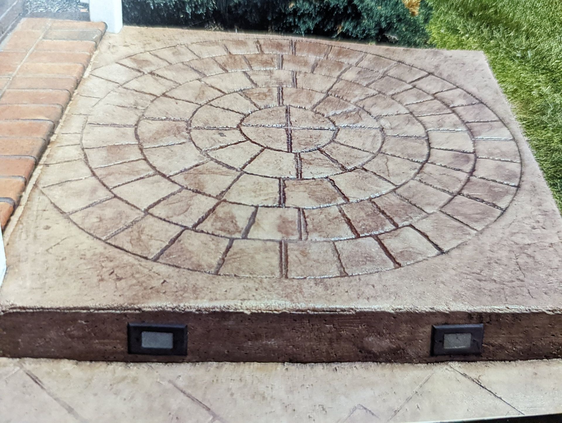 Square concrete utility cover with a circular stone pattern set in a paved sidewalk.