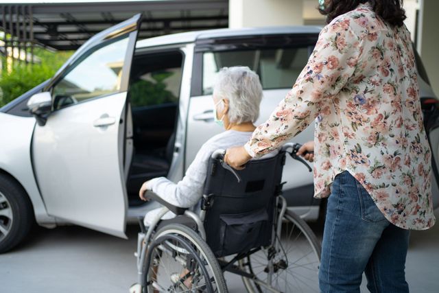 A person in a wheelchair is being assisted by another individual toward an open car door in a driveway.