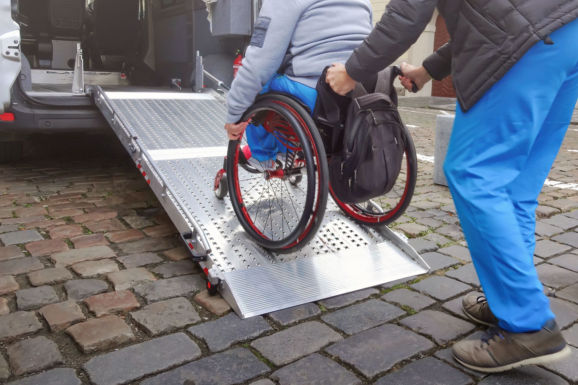 A person pushing a wheelchair user up a metal ramp into the back of a vehicle parked on a cobblestone street. A person pushing a wheelchair user up a metal ramp into the back of a vehicle parked on a cobblestone street.
