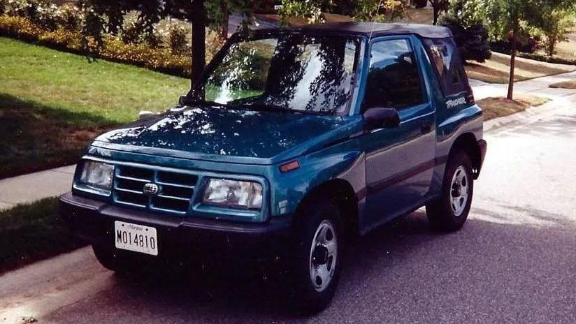 Teal Geo Tracker SUV parked on a paved road next to a grassy hill.