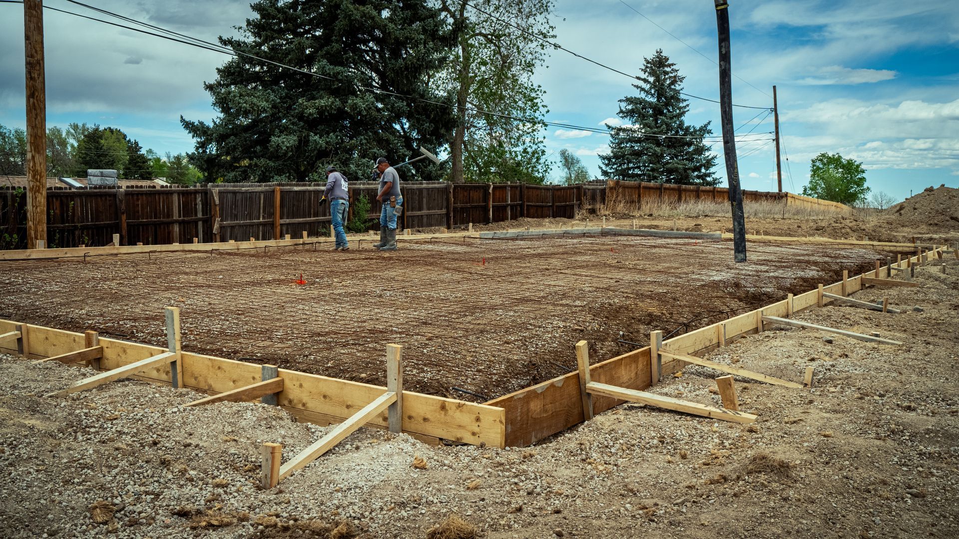Construction site with wooden foundation forms and two workers on a dirt lot under cloudy sky