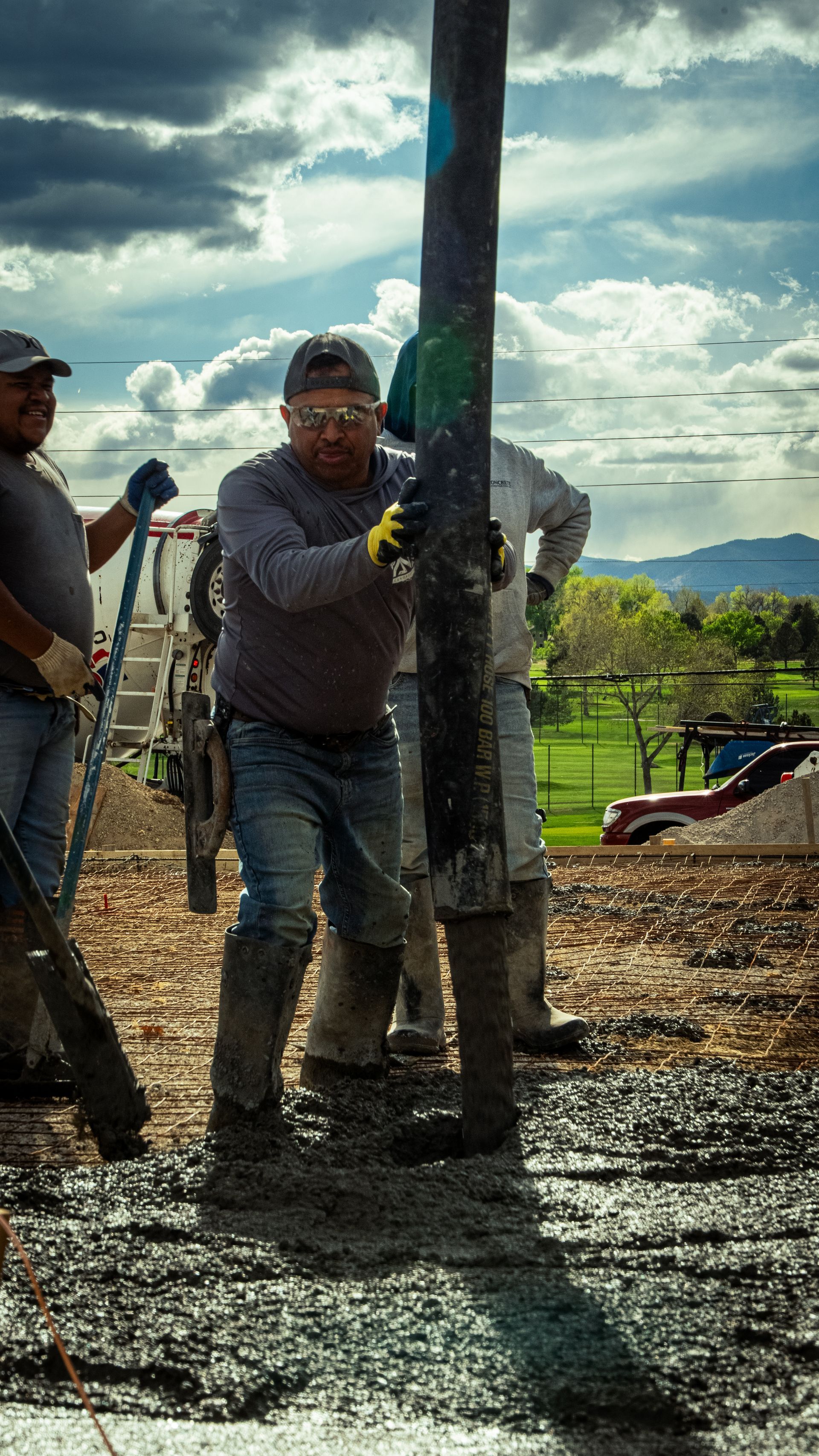 Workers standing in muddy ground lifting a tall pole at a rural construction site under cloudy skies