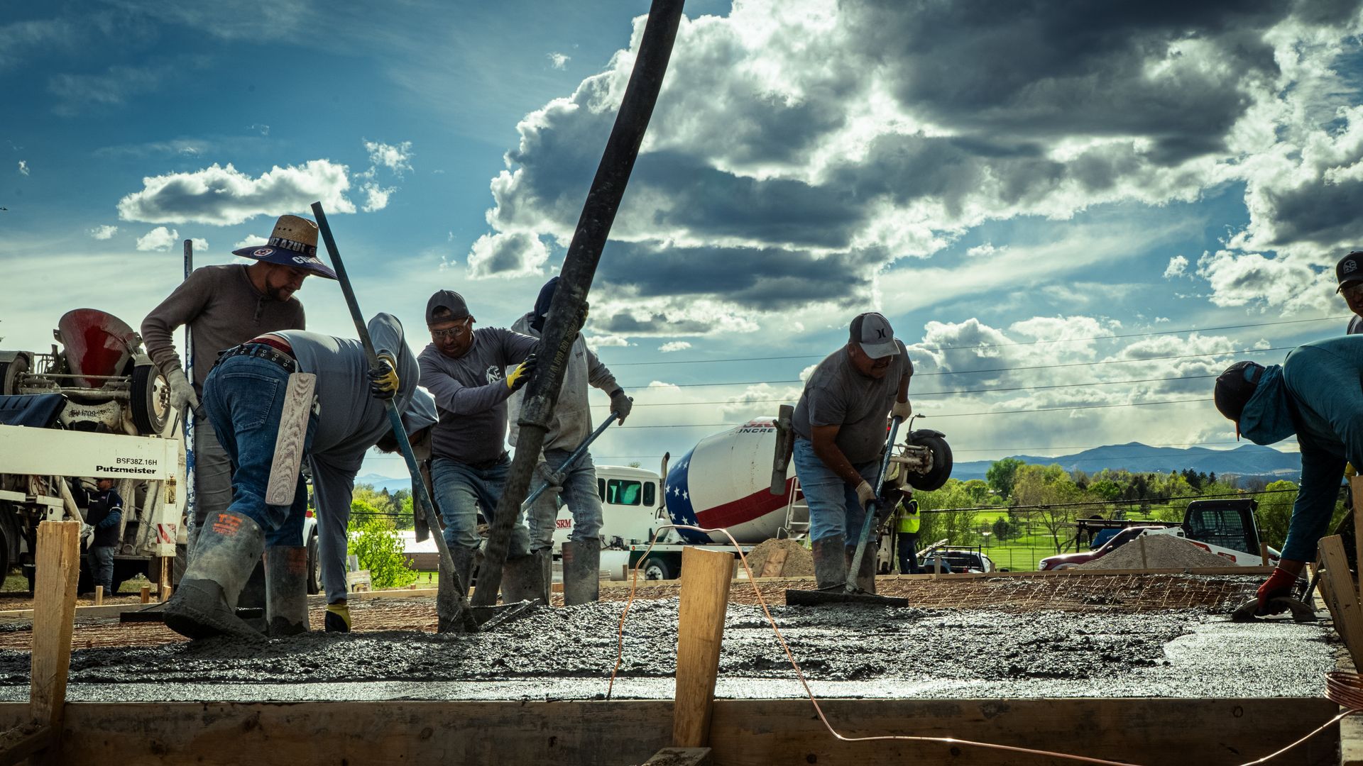 Workers pour concrete at a construction site under a cloudy sky, with a cement mixer and tools nearby.