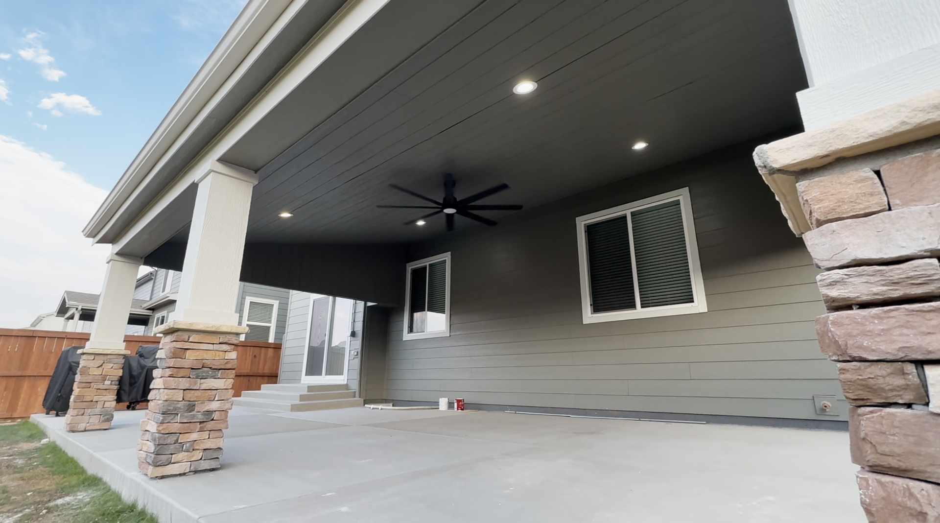 Covered front porch with white columns, stone accents, ceiling fan, and gray siding under a house roof.