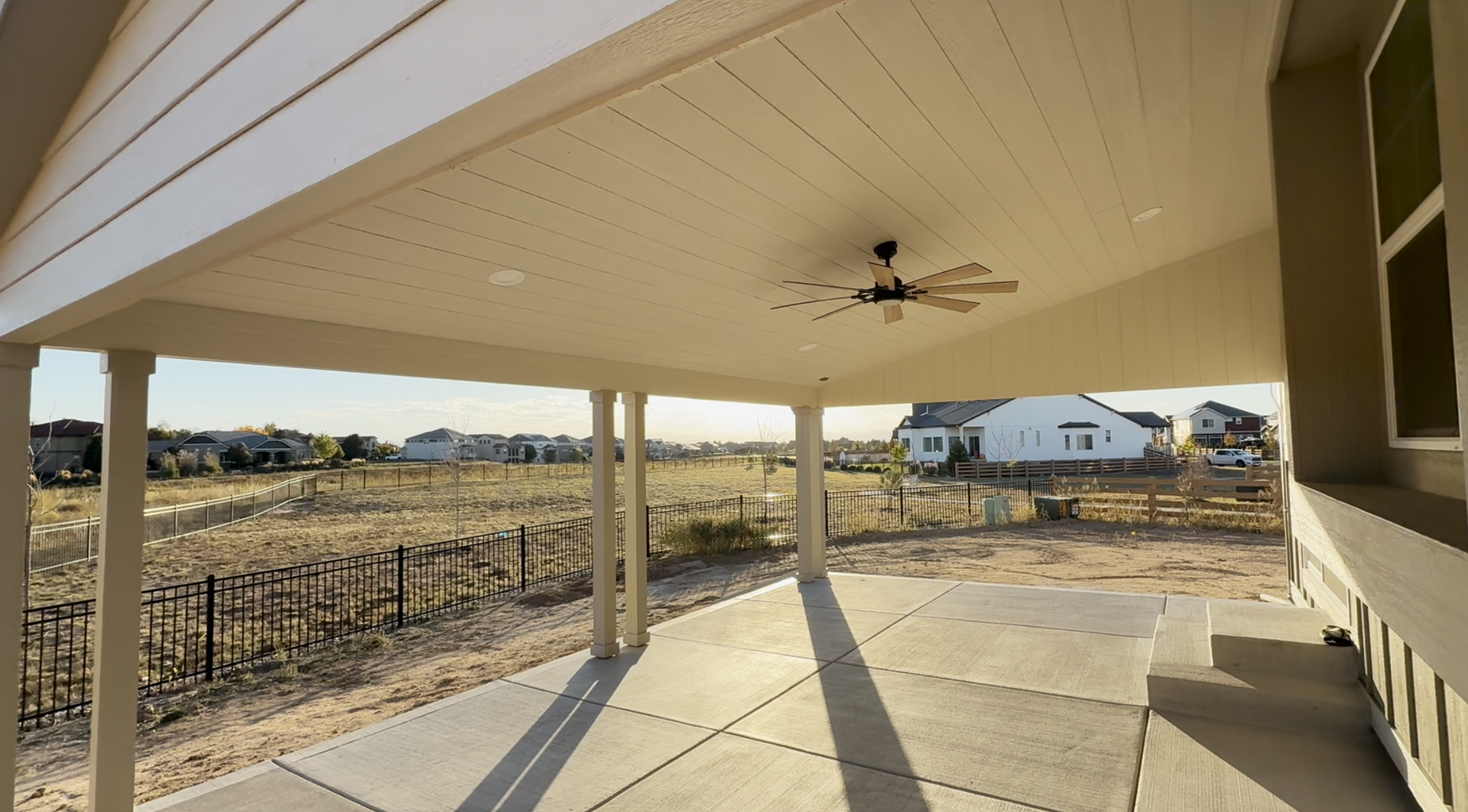 Covered backyard patio with ceiling fan, concrete floor, and view of open fields and neighboring houses