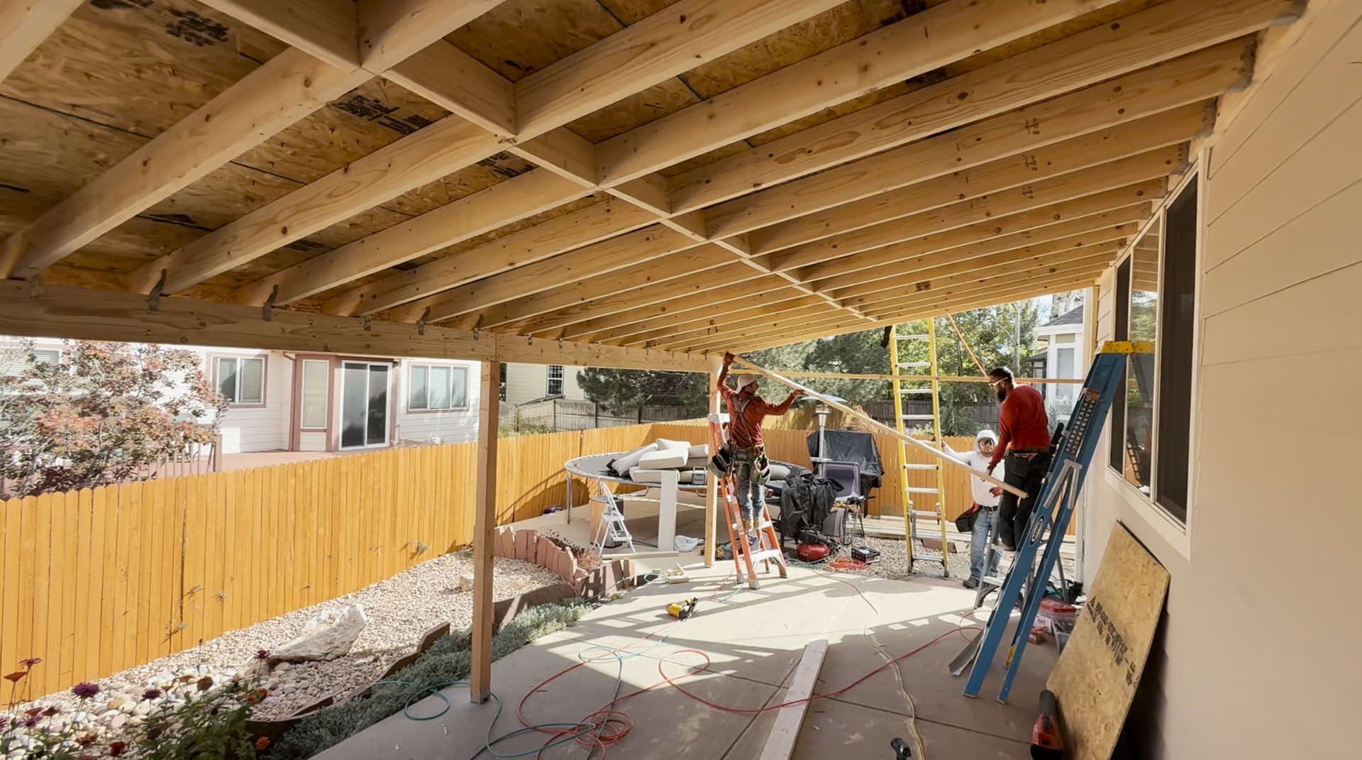 Workers building a covered patio beside a house, with ladders, tools, and construction materials.