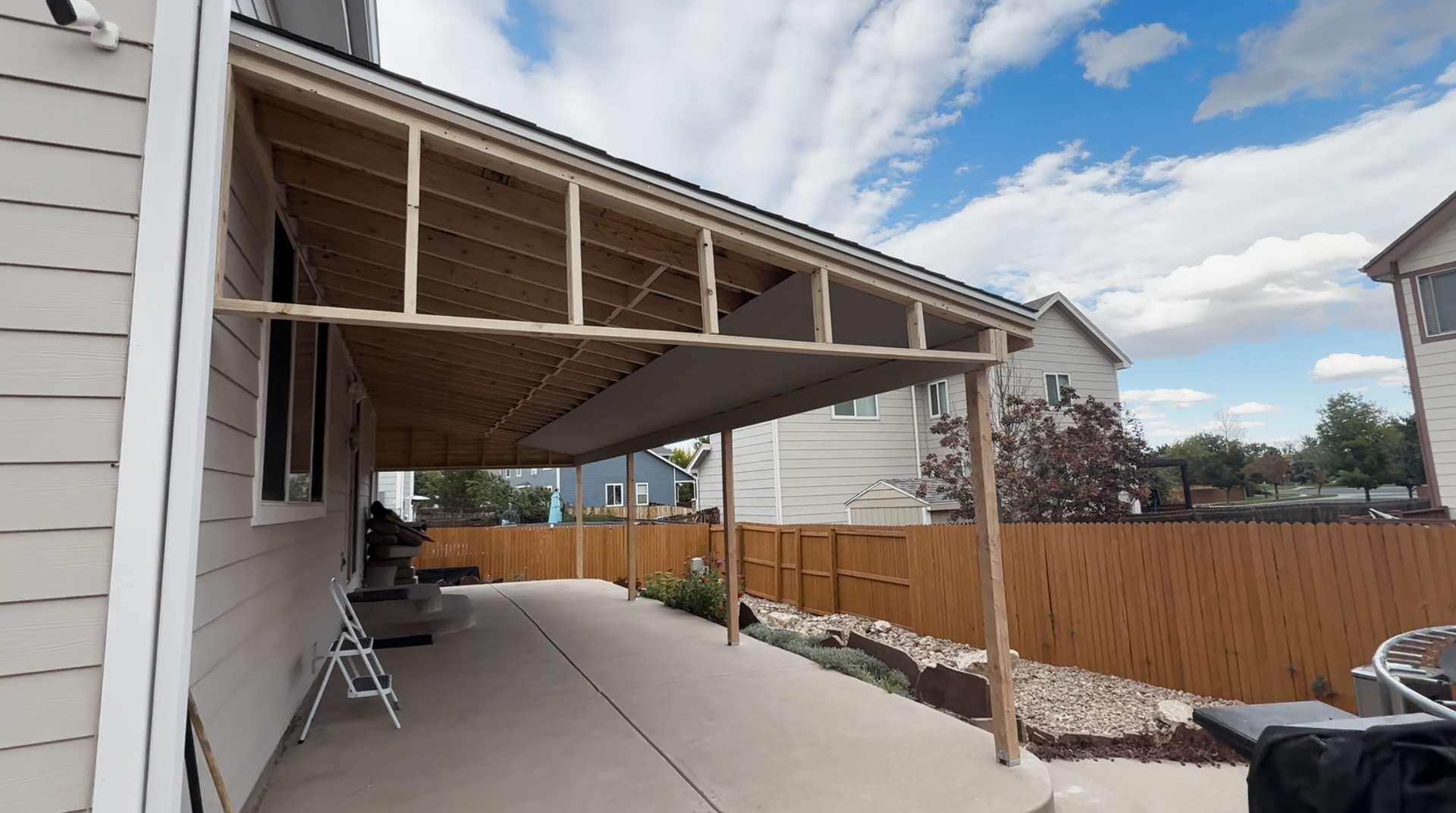 Covered patio beside a house, with concrete slab, support posts, chairs, and a wooden fence under cloudy skies.