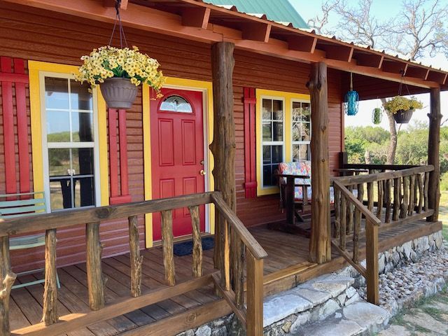 Red cabin porch with wooden railing, yellow-trimmed windows, and a red door with hanging flowers.