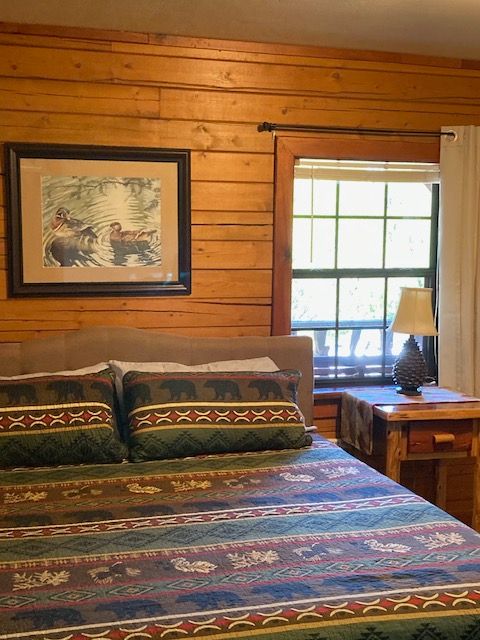 Cozy log cabin bedroom with patterned bedspread, wooden walls, framed art, and a window with a lamp on the nightstand