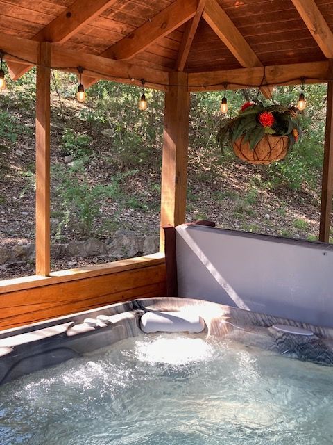 Wooden hot tub under a gazebo with bubbling water and a hanging flower basket, surrounded by trees.