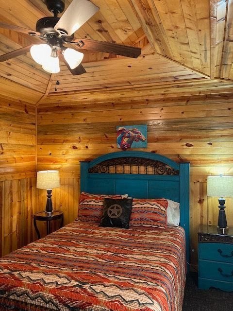 Cozy log cabin bedroom with a teal bed, patterned quilt, lamps, and wood-paneled ceiling with a fan
