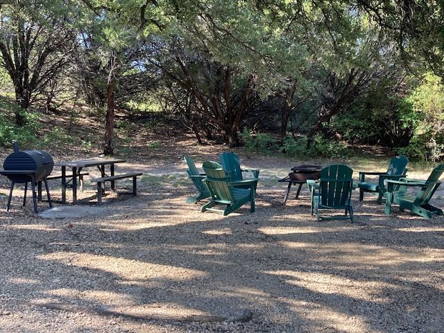 Outdoor picnic area with green chairs, tables, and a grill under trees on a gravel clearing