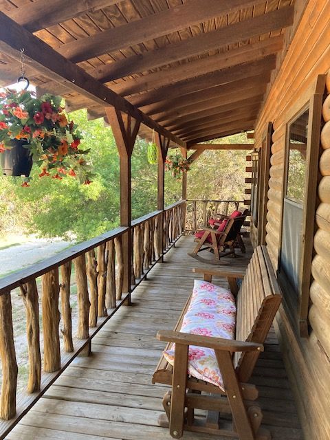 Wooden cabin porch with railings, a bench, hanging flowers, and a red chair overlooking greenery