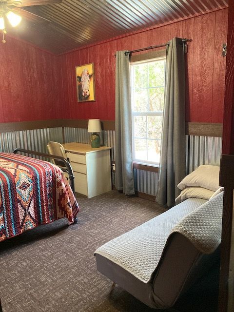 Cozy bedroom with red wood walls, patterned bedspread, window, dresser, and chaise lounge.