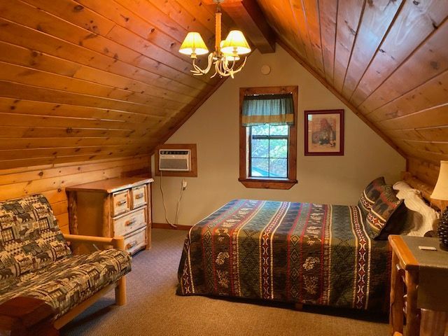 Cozy attic bedroom with wood-paneled walls, a bed, loveseat, window, and warm chandelier lighting.