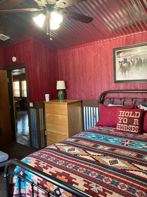 Cozy bedroom with red walls, patterned bedspread, black metal bed, dresser, lamp, and ceiling fan.